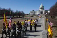 Two rows and two clumns of people march on the Minnesota state capitol grounds carrying wreaths and flags. The capitol rises in the background against a blue sky.